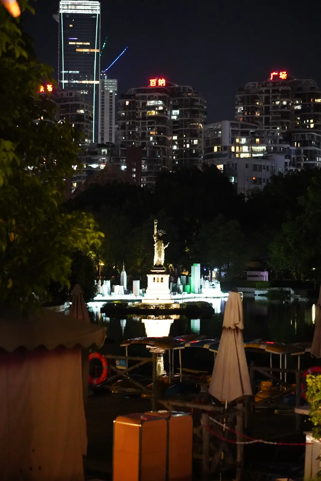 A miniature Statue of Liberty lit across the park lake, city towers behind