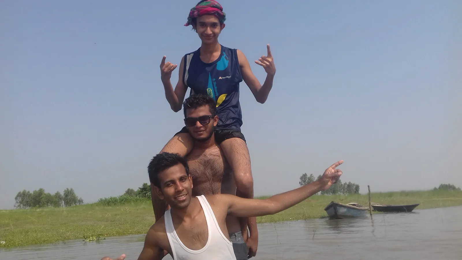 A three-friend human tower in the river, boats on the bank behind